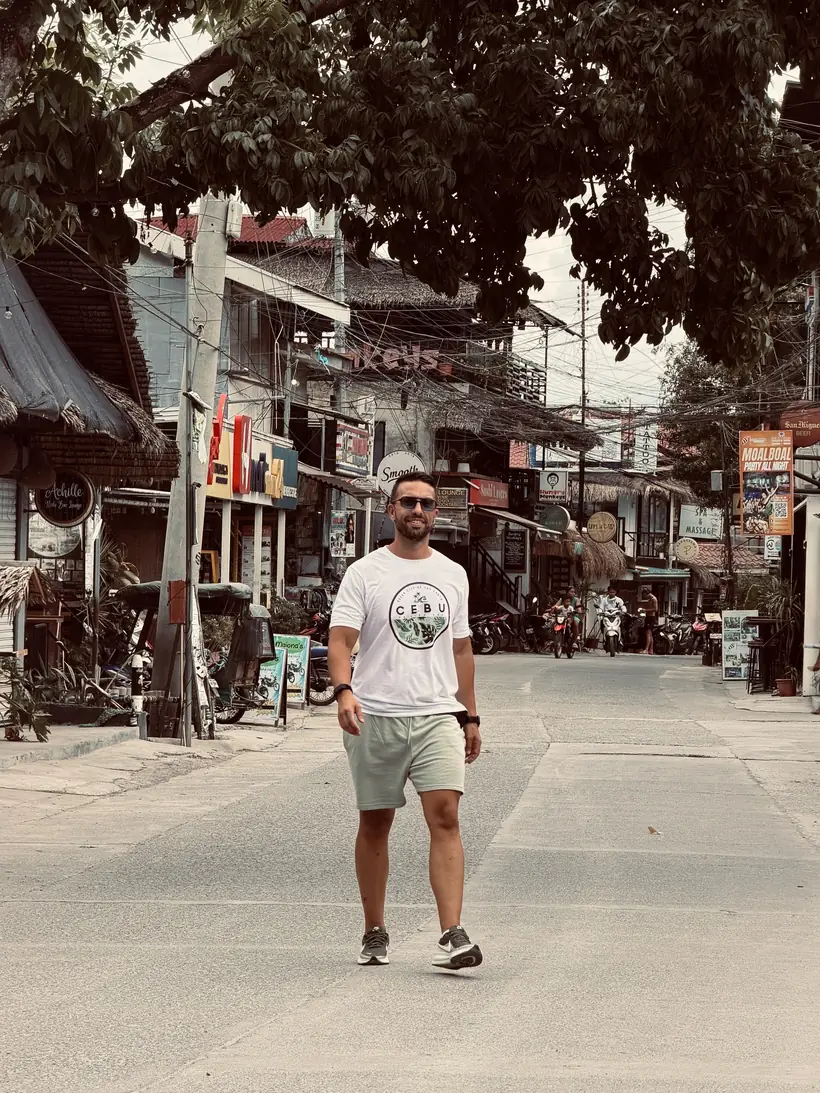 Juan caminando por una calle con tiendas y árboles