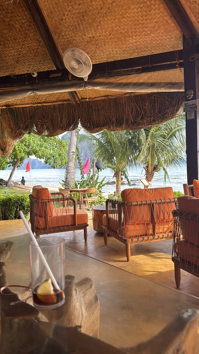 Terraza con sillones naranjas bajo un techo de paja, con una bebida en primer plano y el mar al fondo