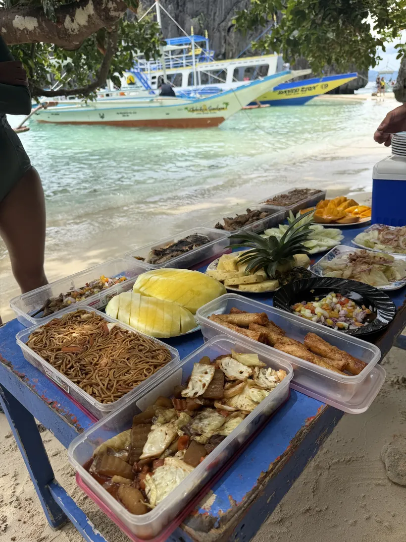 Mesa en la playa con fideos, rollitos, piña, mango y otros platos, con las bangkas al fondo