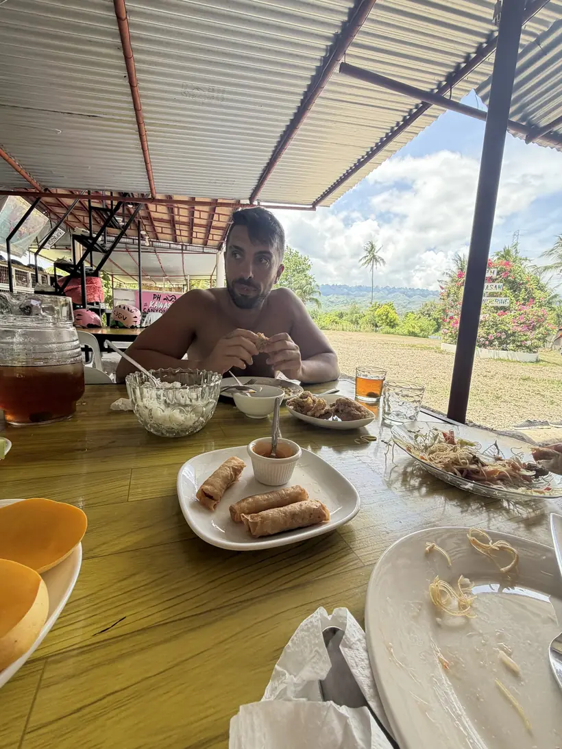 Juan a la mesa con rollitos y otros platos caseros, bajo un tejado de chapa